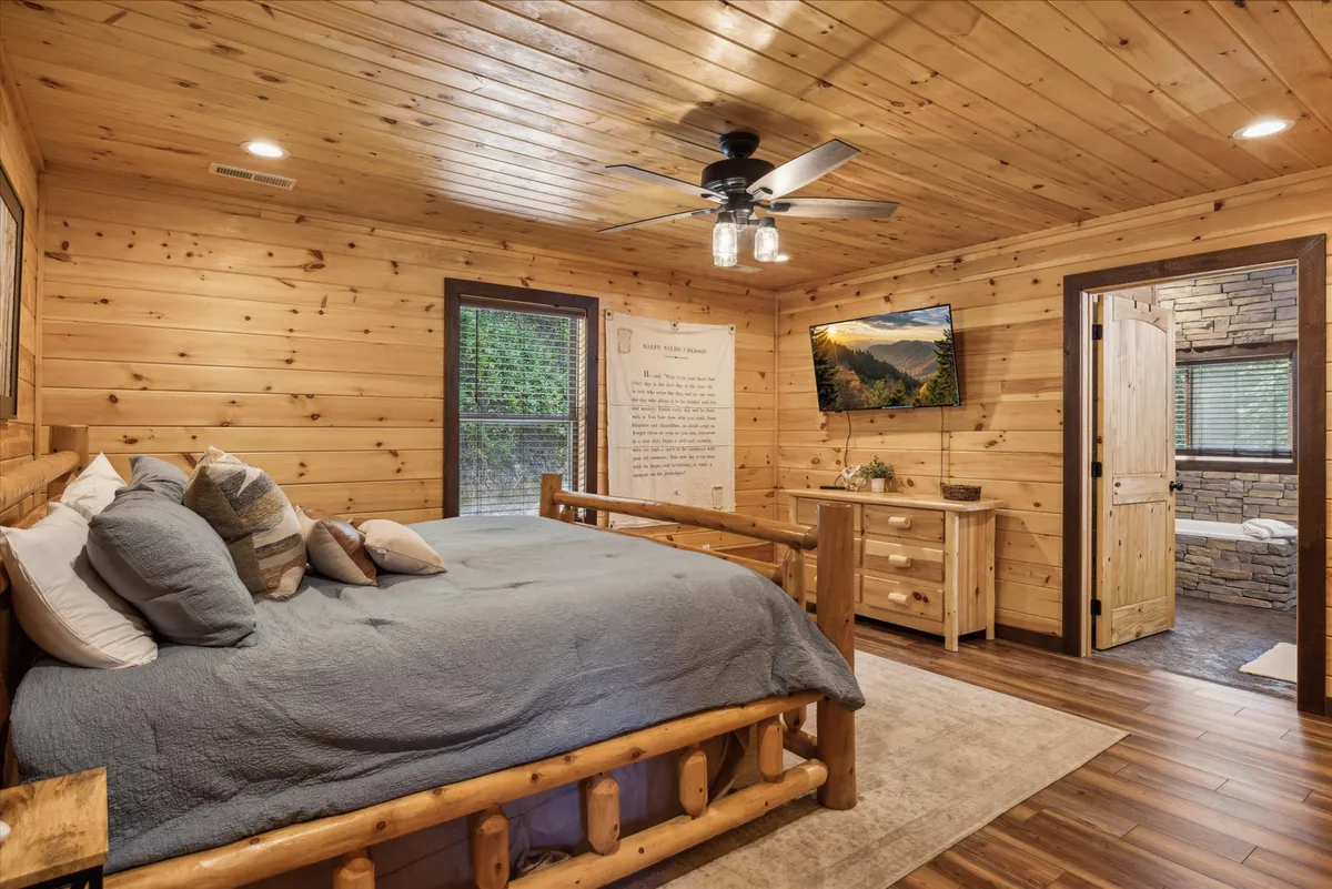 This interior image displays a bedroom featuring a log bed, large window streaming with natural light, framed artwork, a flat-screen TV, and a wooden dresser. The room exudes tranquility with its warm wooden accents and inviting bedding, while the expansive window showcases woodland views.