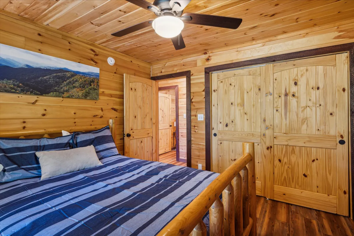 A serene bedroom interior with a bed dressed in striped bedding, wood-paneled walls, a ceiling fan, and nature-themed artwork. The room feels peaceful and cozy, ideal for relaxation or enjoying a quiet morning.