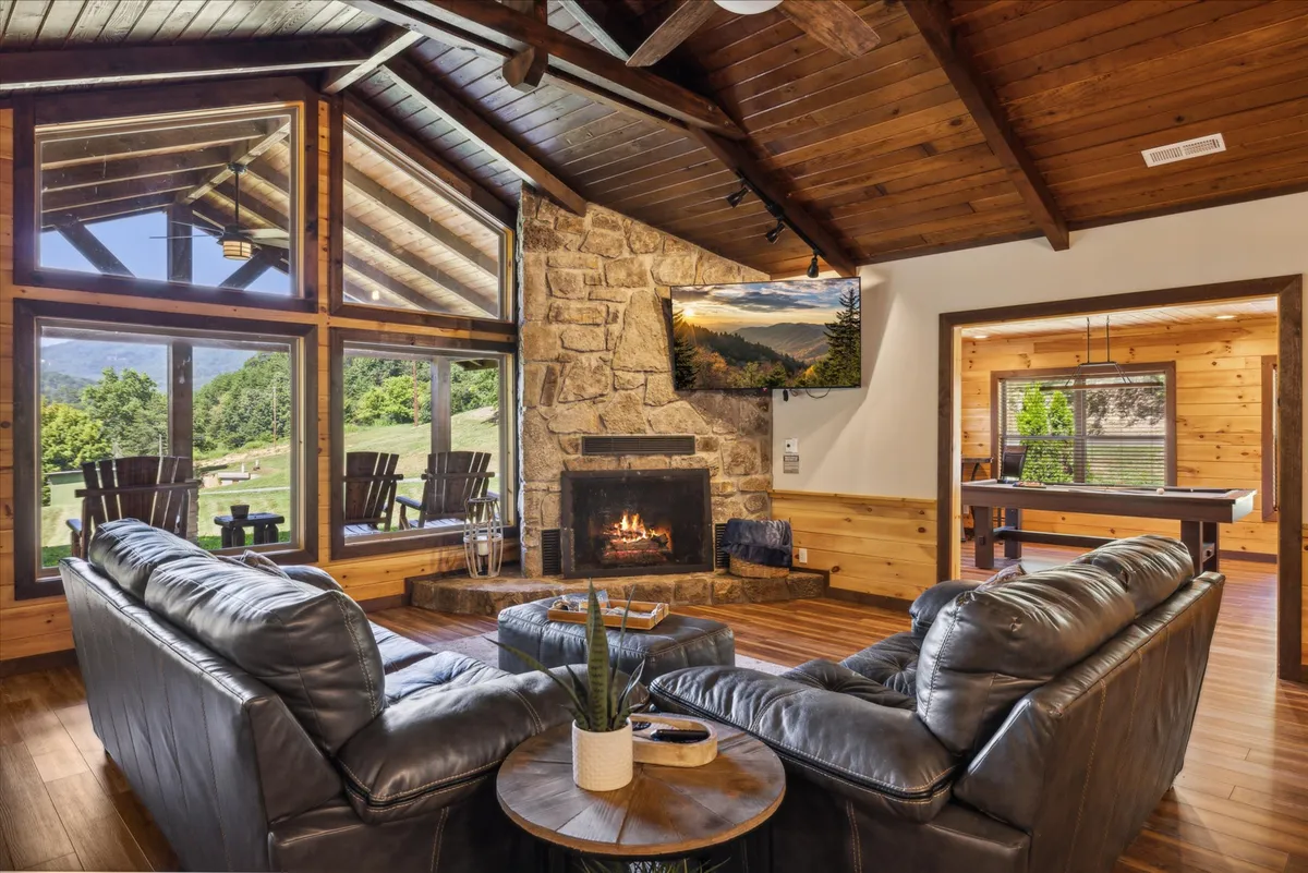 A vibrant living area featuring a comfortable seating group facing a grand, rustic stone fireplace. Large windows offer views of the peaceful landscape outside, while a pool table in the background adds an element of fun and entertainment. The setting is both inviting and versatile, ideal for relaxing, socializing, or enjoying a game night.