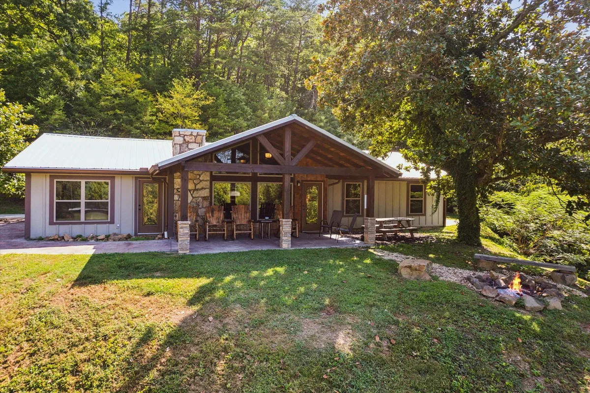 The image showcases the cabin's inviting front porch with classic wooden rocking chairs, a rustic stone fireplace, large windows reflecting the surrounding lush greenery, and expansive views of nature. The peaceful setting hints at unforgettable sunsets, cozy gatherings around the fire pit, and plenty of space to relax and enjoy the outdoors.