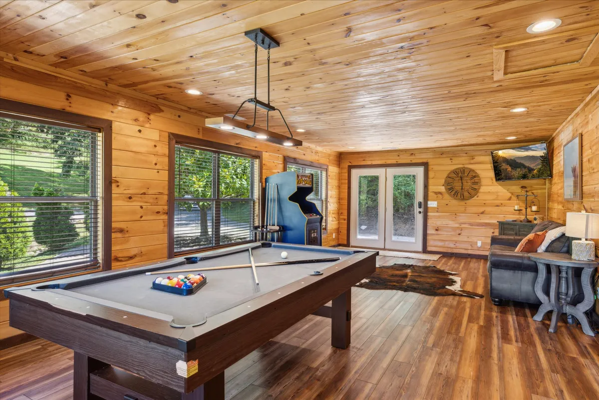A vibrant game room featuring a pool table in the center, a classic Pac-Man arcade machine, a cozy seating area, and expansive windows that let in natural light. The relaxed, playful mood is enhanced by the inviting arrangement, making it perfect for friendly competitions or laid-back lounging.