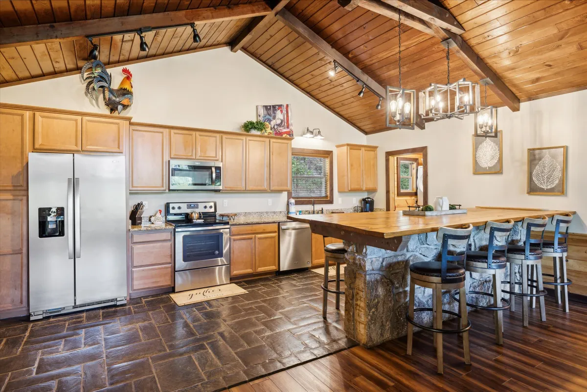 A cozy kitchen scene with a large wooden island accented by a stone base, stainless steel appliances, and rustic wooden beams overhead. The decor includes a decorative rooster, adding a touch of hominess. The kitchen feels welcoming and designed for both entertaining and meal preparation, echoing the social hub described in the section.