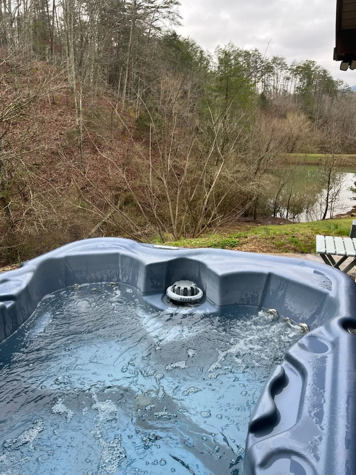 An outdoor hot tub nestled in serene natural surroundings with a calm water body nearby. The hot tub is the focal point, inviting guests to unwind while enjoying views of the surrounding greenery, creating a sense of privacy and relaxation.