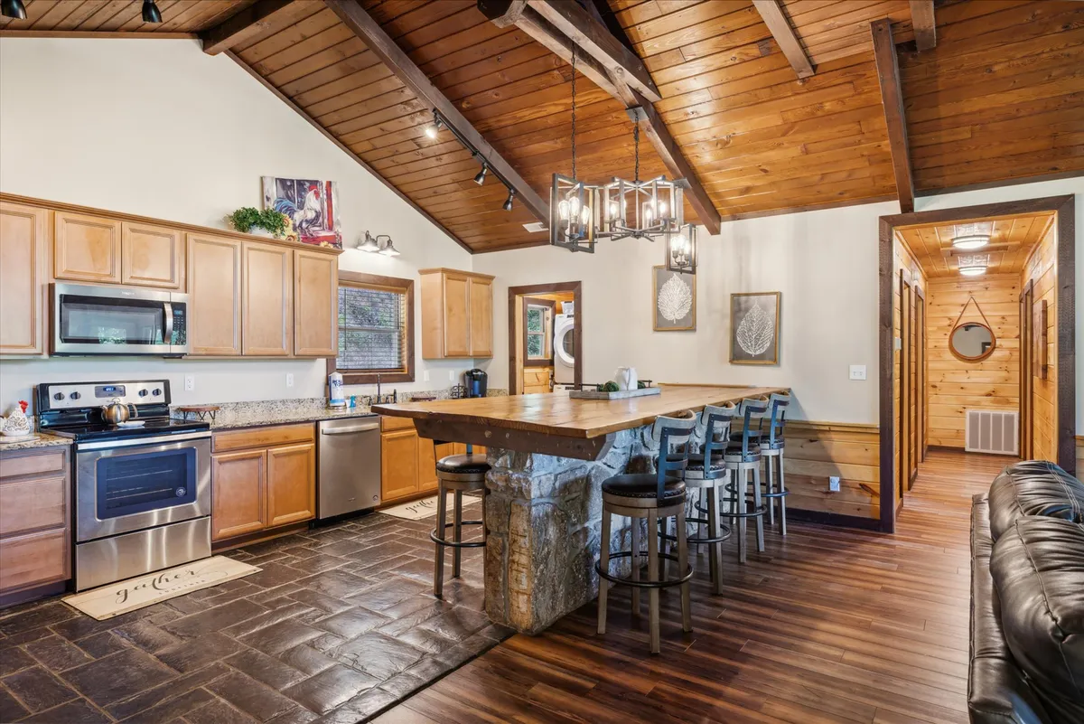 This image features a spacious, fully-equipped kitchen with a large wooden kitchen island that has a striking stone base. Stainless steel appliances, ample wooden cabinetry, and a high ceiling with exposed beams create a warm yet modern atmosphere. The open layout invites gathering and culinary activity, while the island offers generous counter space for meal preparation or casual conversation.