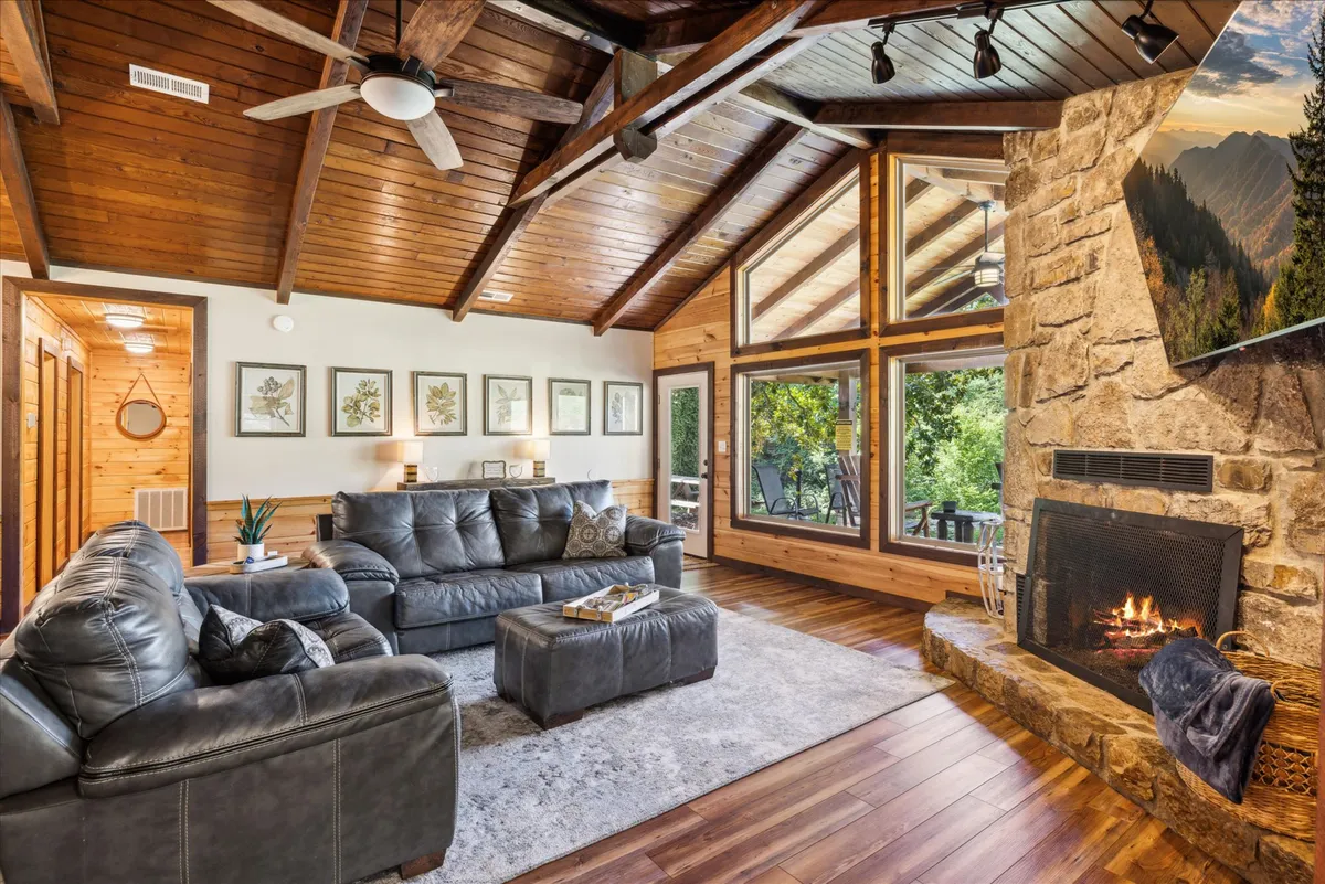 A spacious living room with a leather sectional sofa, a prominent stone fireplace, and large windows that flood the space with sunlight and frame tranquil outdoor views. Exposed wooden beams and warm lighting create an atmosphere of comfort and hospitality, emphasizing the room’s suitability for group relaxation.