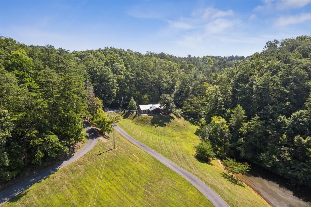This image provides a wider context, showcasing the house nestled among trees with a winding driveway and a scenic river nearby. The tranquil setting emphasizes the home’s seclusion and its natural surroundings, inviting exploration and outdoor adventures just steps away from the front door.