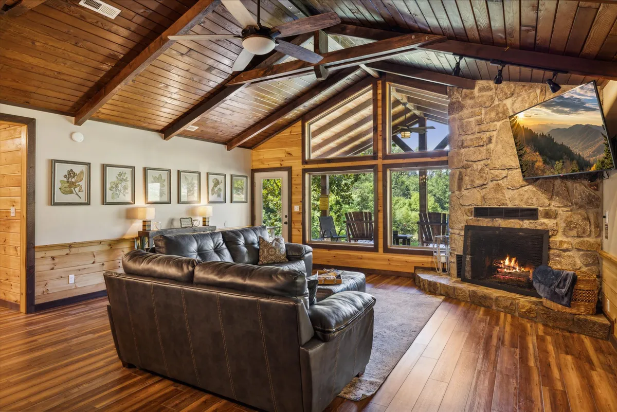 A warm and welcoming living room featuring a large leather sofa arranged around a striking stone fireplace. Expansive windows let in abundant natural light and offer serene views of the surrounding nature. Wooden beams and ceiling add rustic charm, while the open layout creates an inviting gathering space perfect for socializing or relaxing.